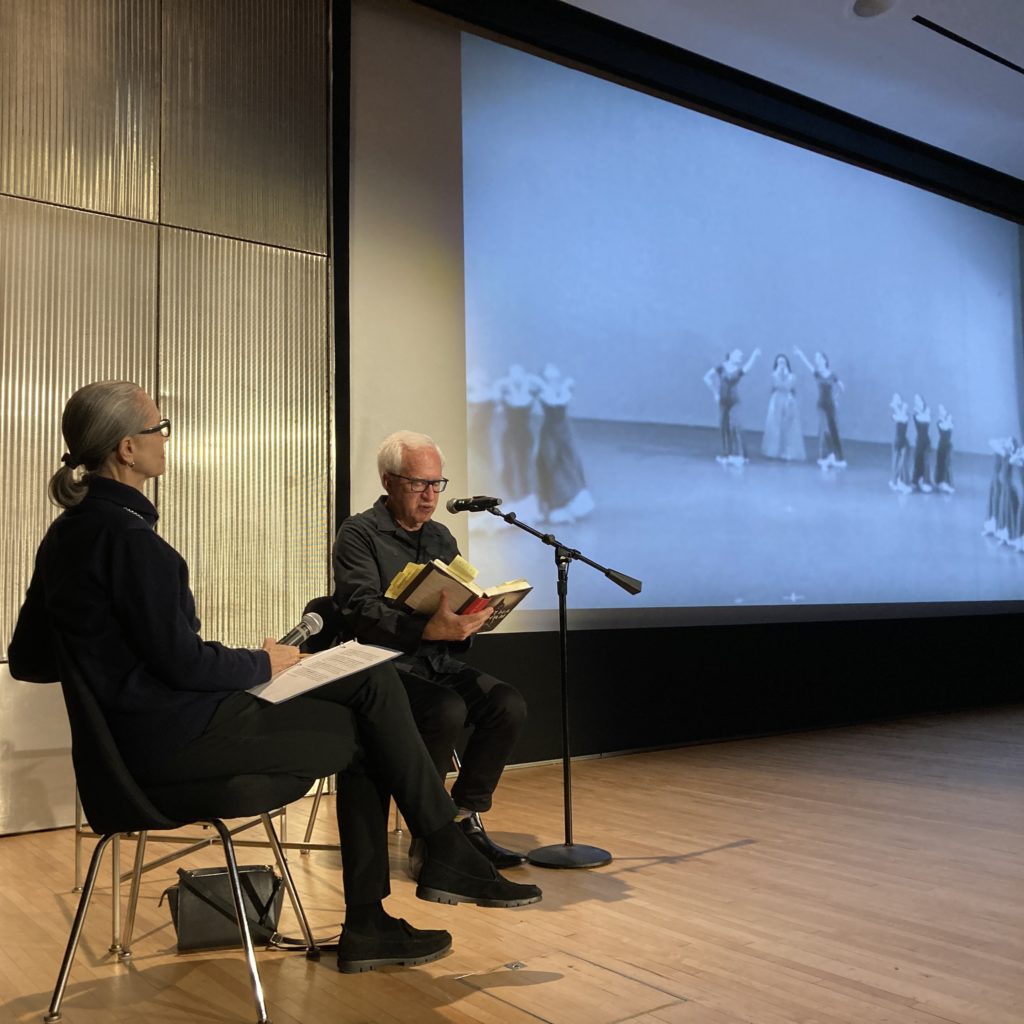 A woman sits with a microphone next to a man reading a book into a microphone. Next to them is a large screen that shows a black and white image of a dance by Martha Graham with four groups of three people.