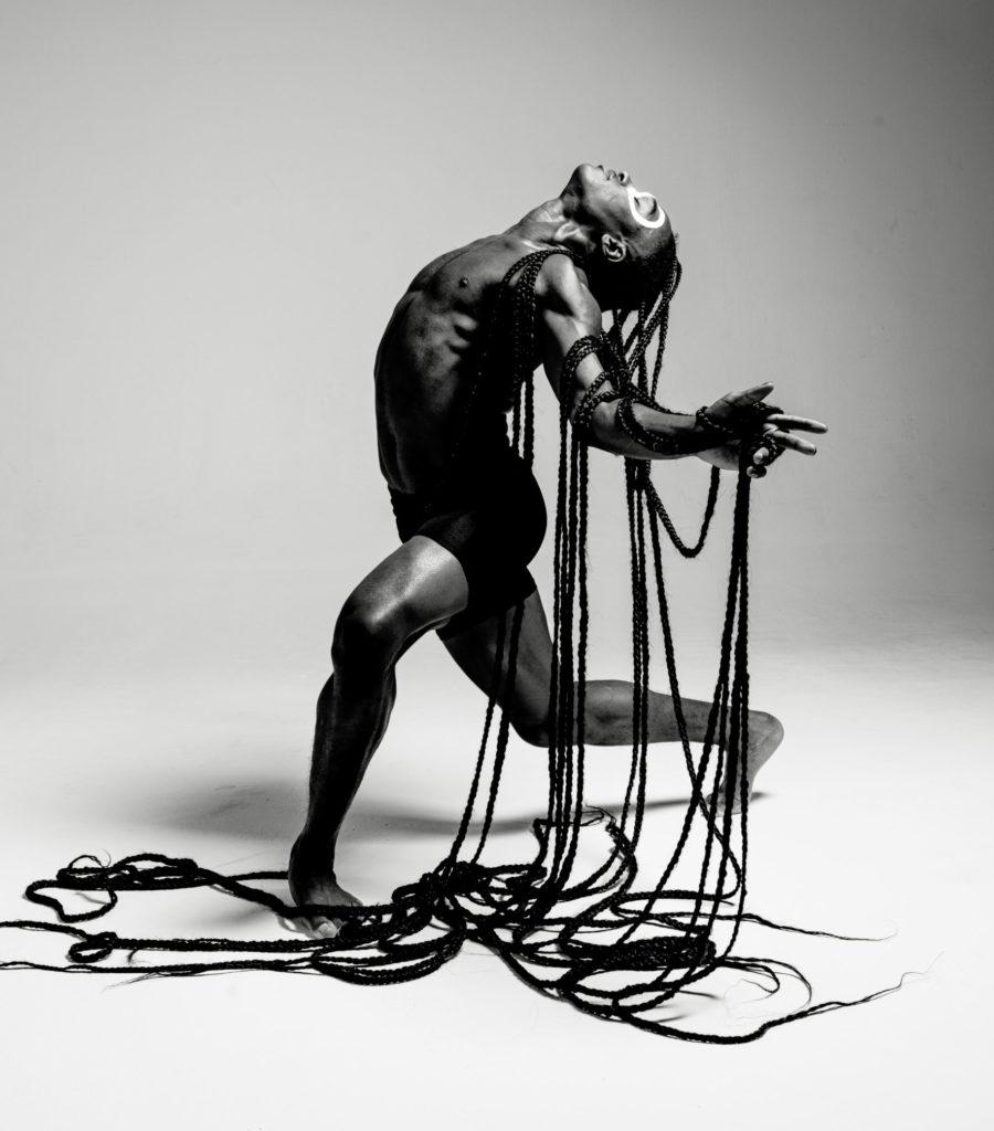 A black-and-white image of Jamal Wade, a Black man arching backwards as he lunges. He has hair extensions several feet long.