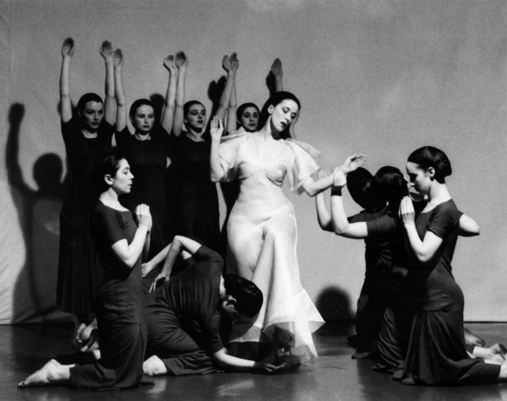 Martha Graham, in a long white dress, stands surrounded by a group of dancers in black dresses, on their knees on each side of her and standing with arms raised behind her.