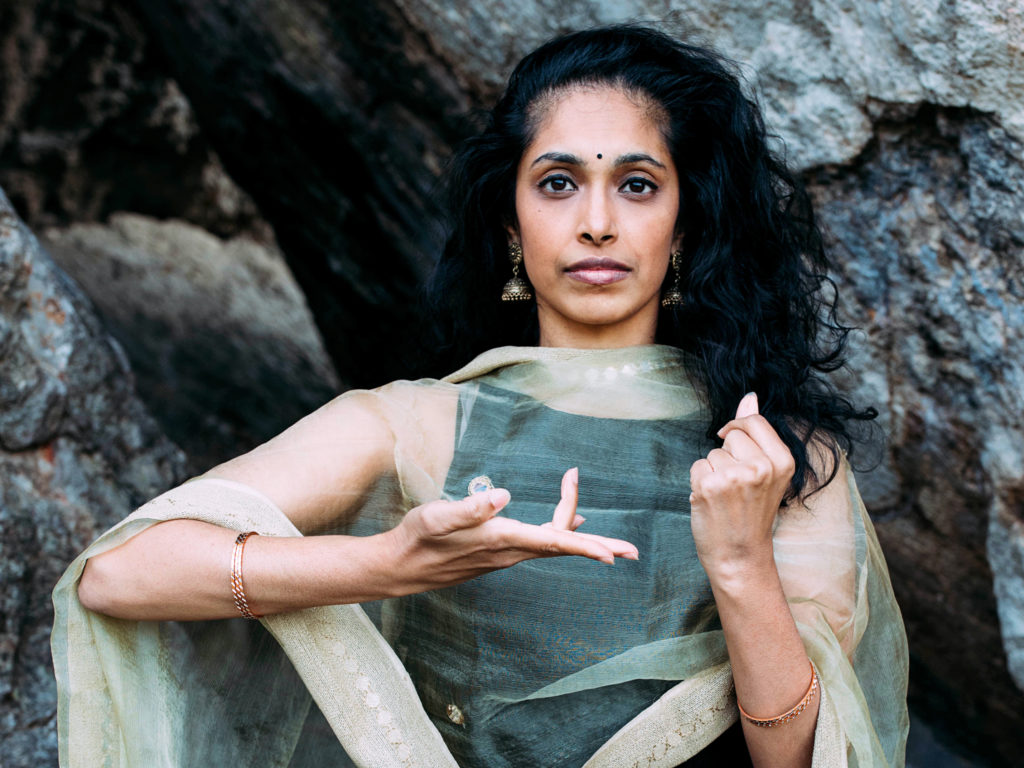 Nadhi Thekkek gazes serenely at the camera. She is a brown skinned woman with her dark hair in loose waves and a dark bindi at the center of her forehead. One of her hands is closed in a fist, palm toward her chest; the other seems to gesture, palm up, toward it, her fourth finger and pink curling lightly upward.