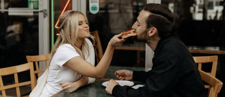 A woman in a white dress sits at a table with a man in all black, holding a slice of pizza that he takes a bite of.