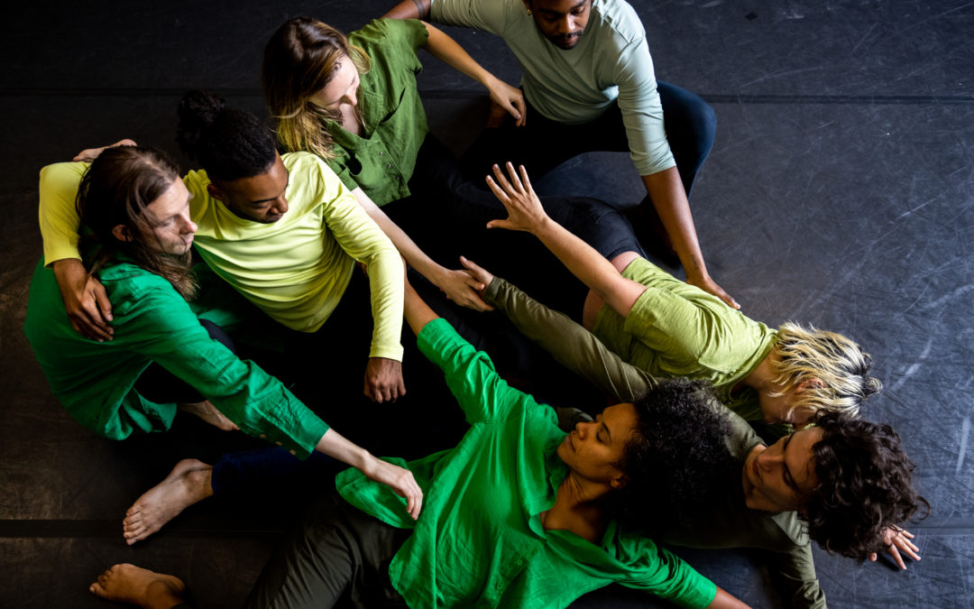 Seven interconnected dancers sprawl and crouch on a dark marley floor. Three who are tipped onto their sides reach their arms toward the loose, clustered line of the other four. All wear shirts in shades of green and dark bottoms.