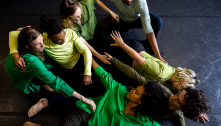 Seven interconnected dancers sprawl and crouch on a dark marley floor. Three who are tipped onto their sides reach their arms toward the loose, clustered line of the other four. All wear shirts in shades of green and dark bottoms.