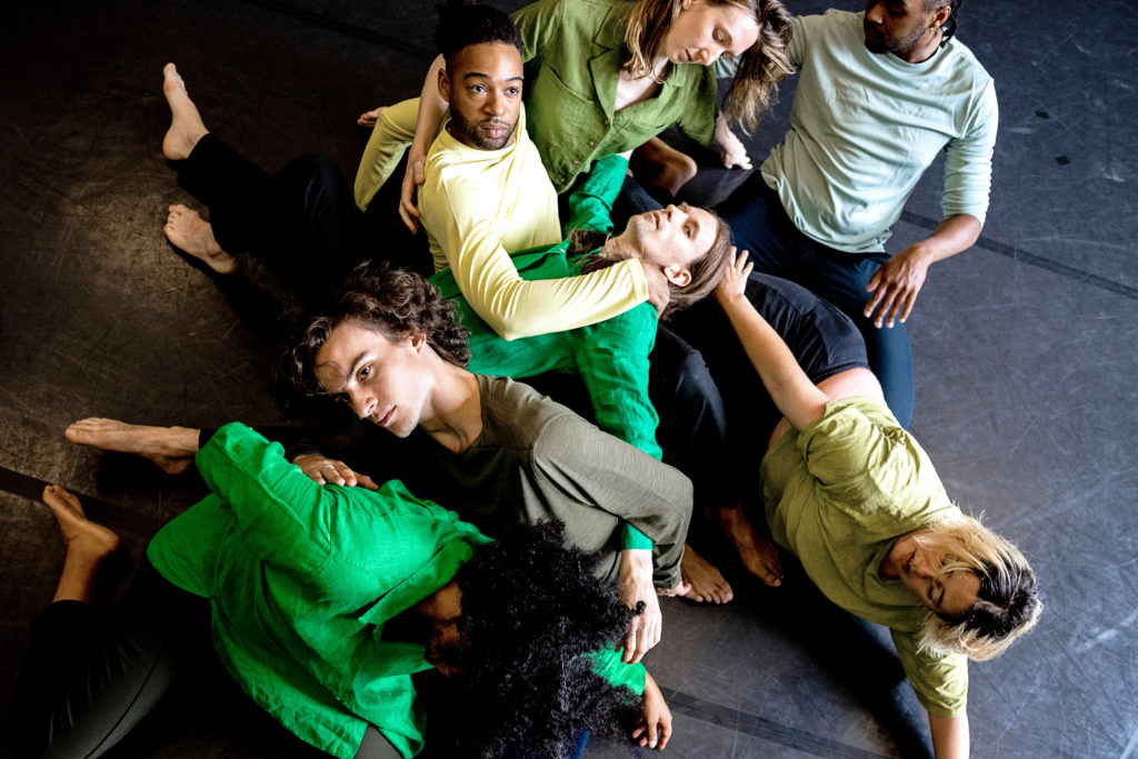 A cluster of seven dancers is shown from above as they cluster and sprawl, interconnected, on a dark marley floor.