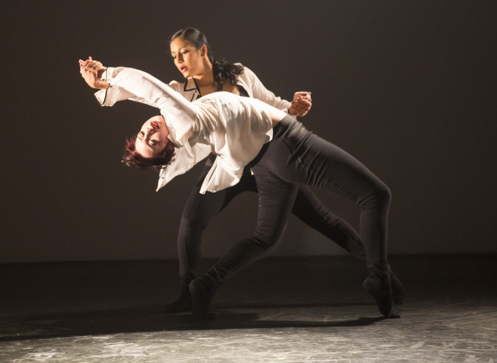 Two women in white shirts and black pants partner each other. One bends backwards on forced arches as the other dancer holds her hands.