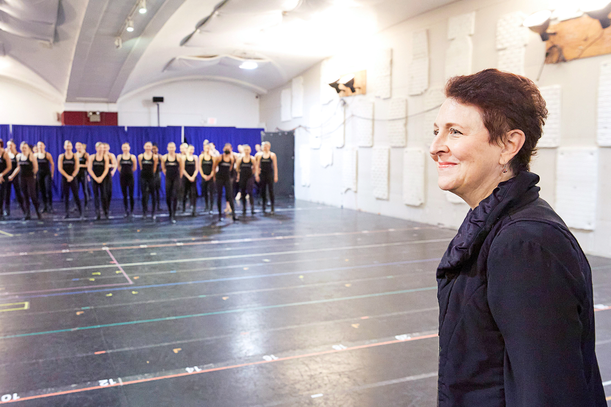 Julie Branam is seen in the foreground, standing in profile with a closed-lip smile. Across the studio, over a dozen women in matching black rehearsal-wear stand to one side.