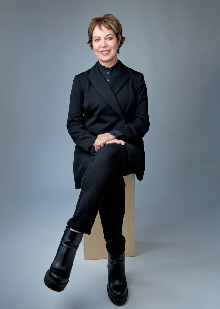 female with short brown hair wearing all black while sitting on a stool