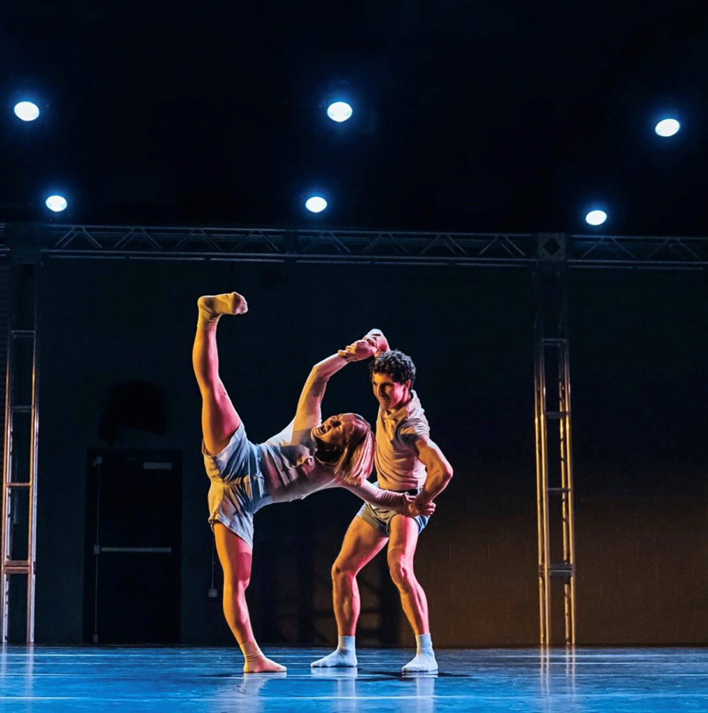 two dancers on stage, male holding females hands while she performs flexed foot tilt