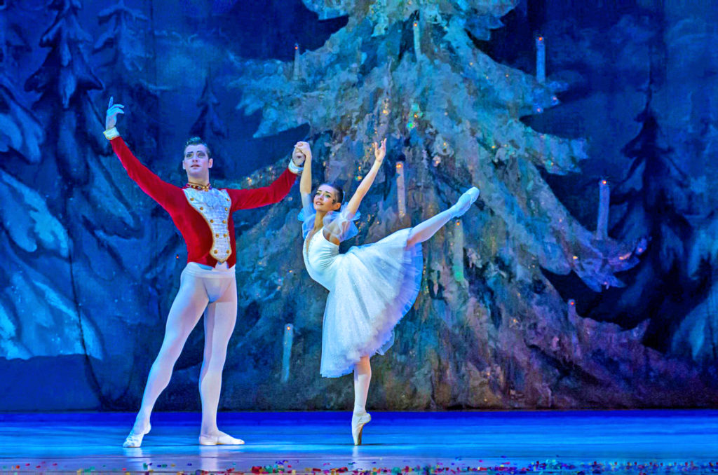 A dancer in a white dress with a knee-length tulle skirt balances in a high arabesque en pointe, a male partner wearing a red tunic helping her balance with one hand holding hers. A Christmas tree and a painted set of snow-dusted evergreens are in the background.
