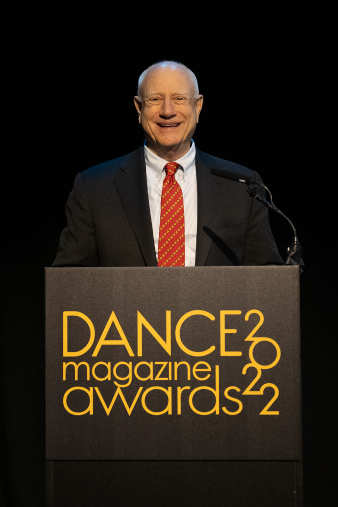 Jim Herbert, an older white man in a suit with a red tie, stands smiling behind a podium bearing the 2022 Dance Magazine Awards logo.