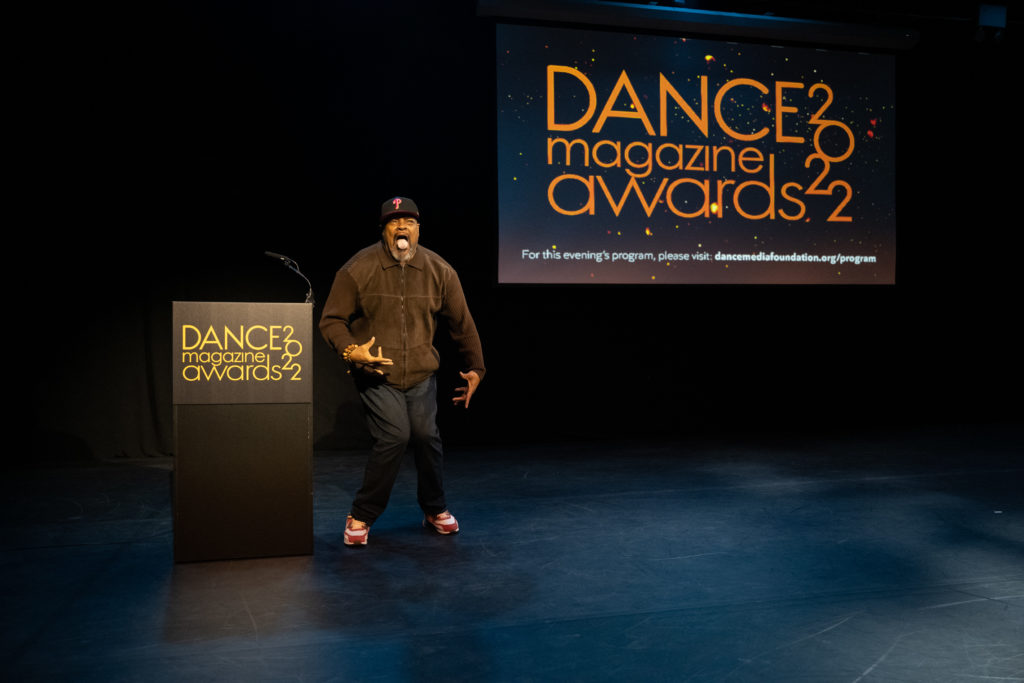 Rennie Harris stands pigeon-toed beside the podium, mouth wide and tongue sticking out, fingers splayed in the air around his torso. A screen at the back of the stage displays the text, "Dance Magazine Awards 2022."