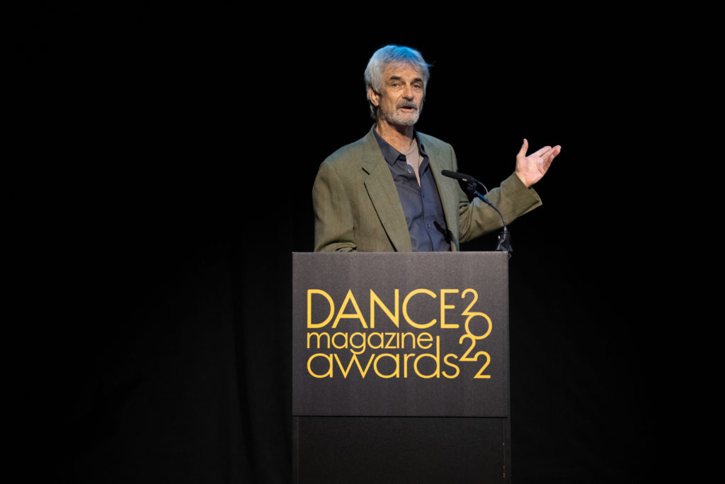 Kevin McKenzie gestures with one hand toward the out-of-frame projection screen, speaking from behind a podium.