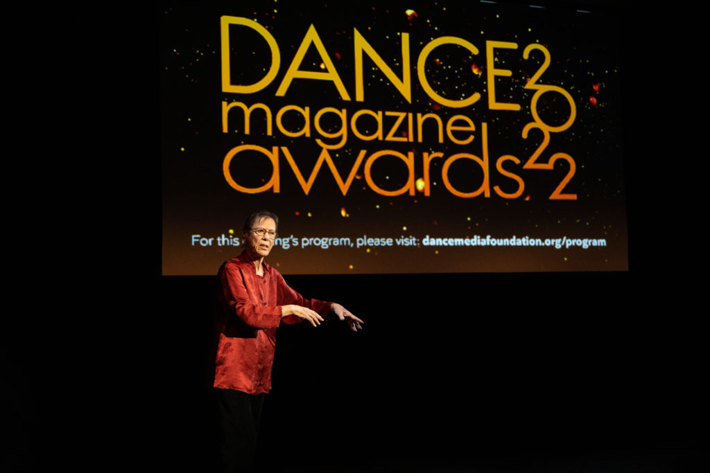 Yvonne Rainer gestures with both hands to one side, illustrating the story she is telling. She has left the podium behind, intent on her demonstration. Behind her, a screen reads, "Dance Magazine Awards 2022."