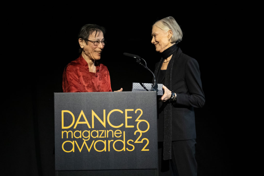 Yvonne Rainer smiles broadly as she hands the box holding a Dance Magazine Award to Lucinda Childs as the two meet behind the podium.