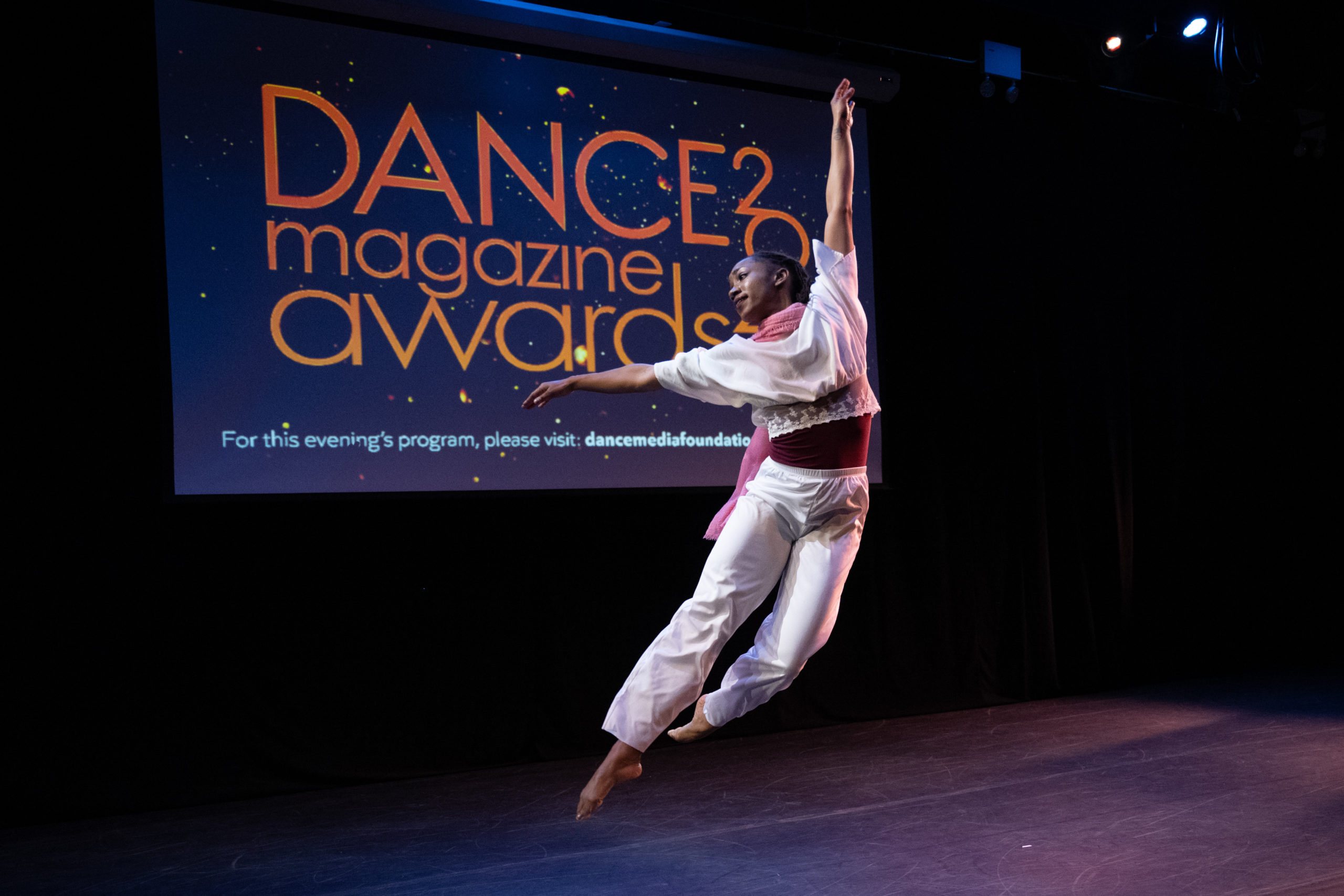 A Black dancer in flowing white pants and a half-sleeved crop top is caught midair in a petite jeté, looking over her side-extended arm in profile. In the background, a projection screen shows the logo for the 2022 Dance Magazine Awards.