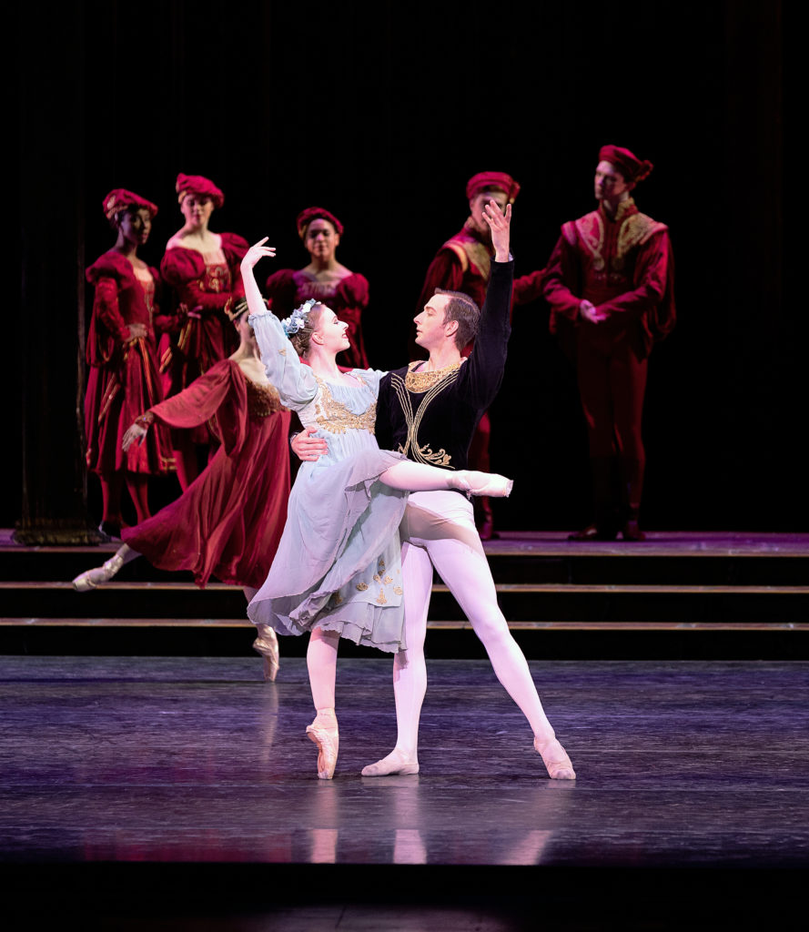 Erin Casale balances in attitude front en pointe, her partner, the prince, supporting her around the waist and mirroring her outside arm in high fifth. She wears a pale blue dress with golden details and finery. Courtiers in red look on from upstage.