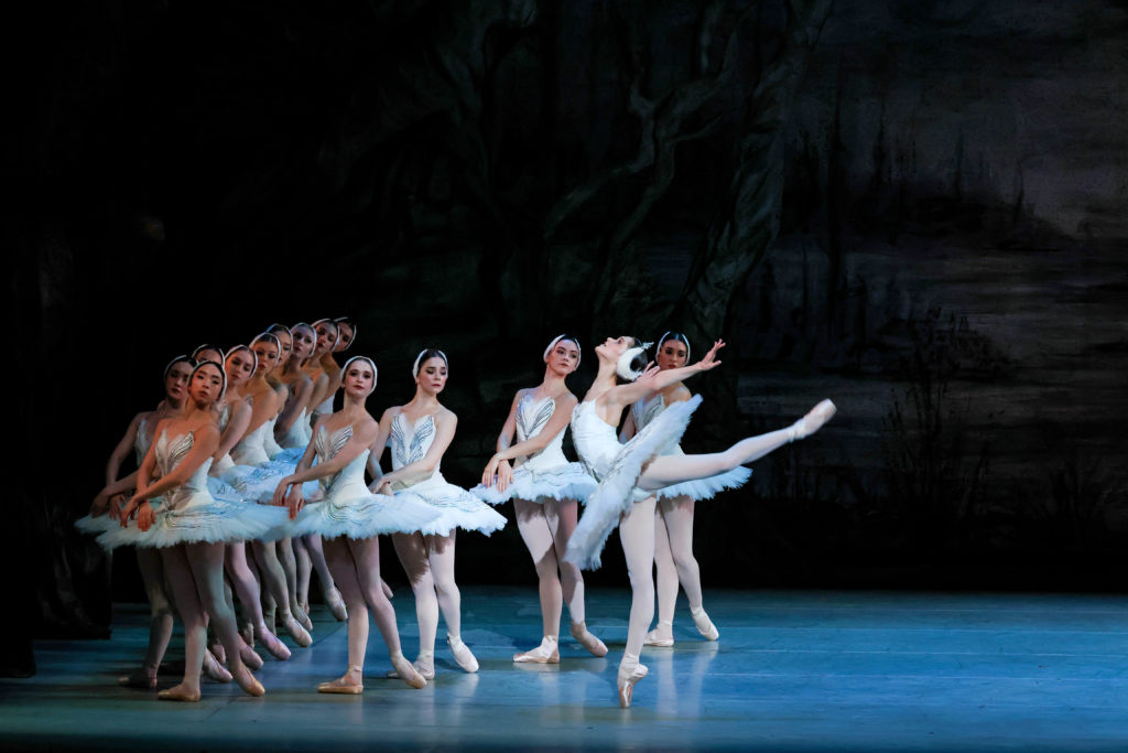 Adelaide Clauss balances in attitude back en pointe, facing the wings, her head tipped to the sky and arms extending wing-like past her shoulders. Over a dozen corps dancers in matching white tutus pose in a V, each standing in an open B-plus, wrists crossed to hover just over their tutus.