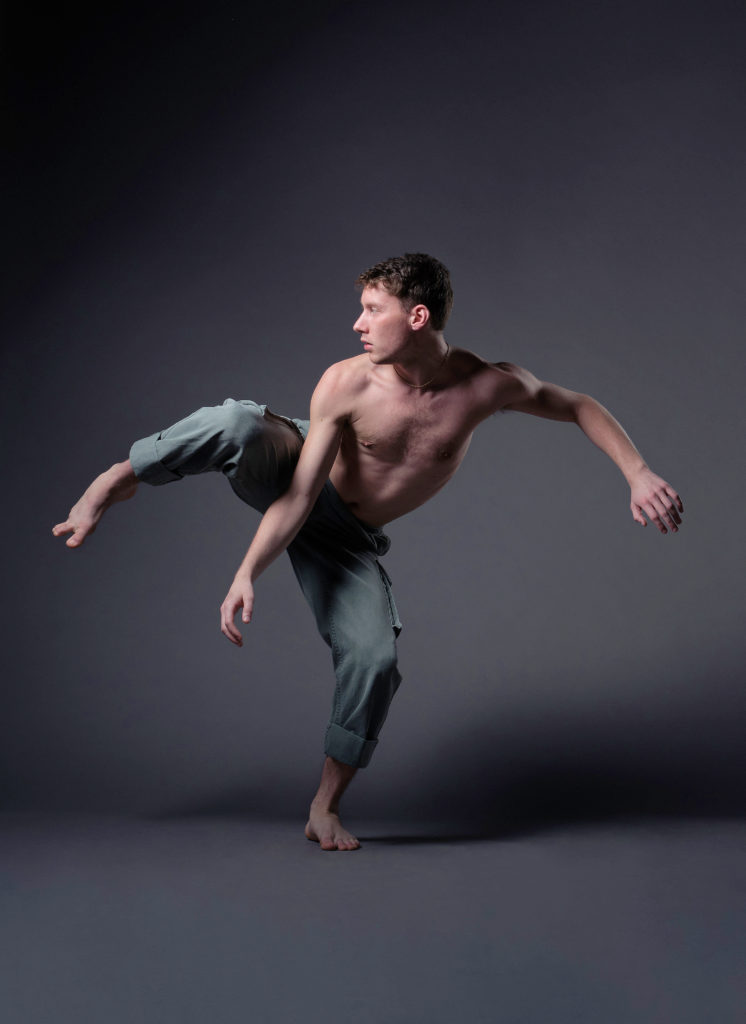 Andrew McShea poses barechested and barefoot in front of a grey backdrop. He looks to his right as his right leg rises in a side attitude, foot arching toward the floor. His opposite arm mirrors his working leg as he hunches slightly forward over his bent standing leg.