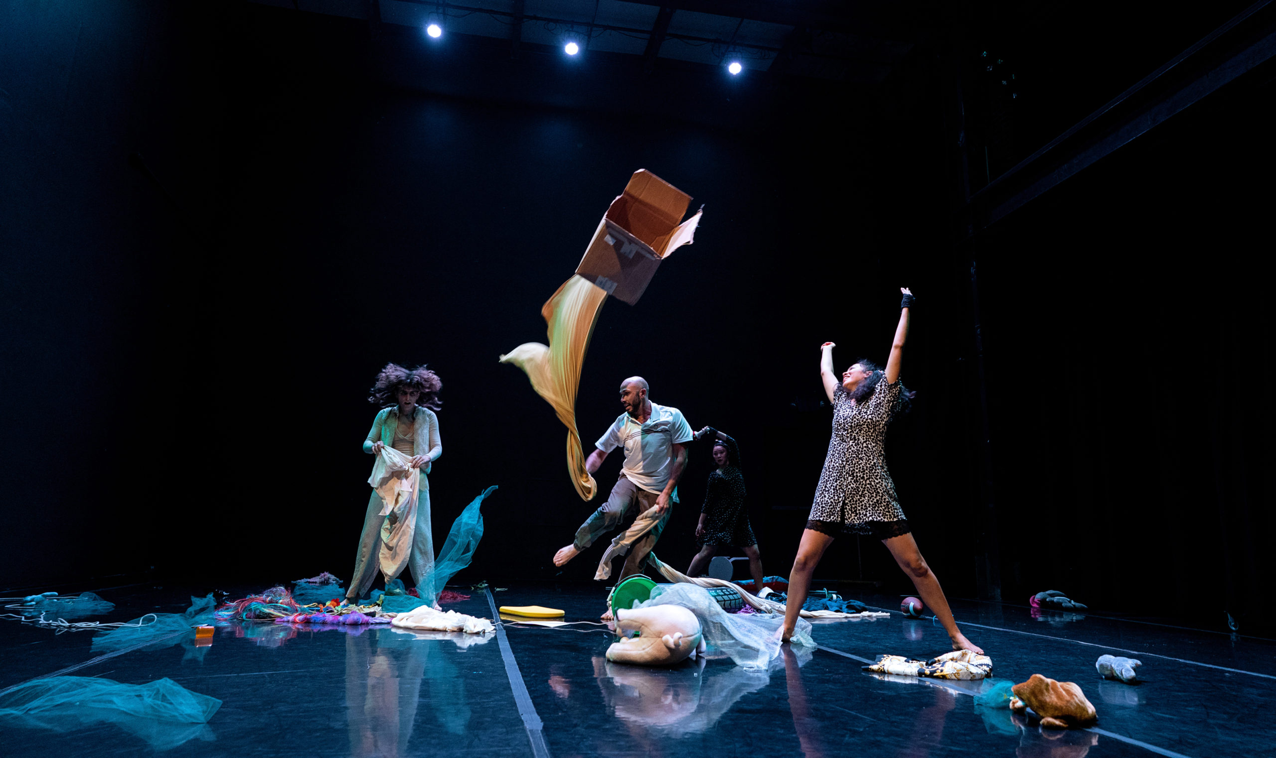 Four dancers variously run and stand on a stage littered with fallen boxes, stuffed animals and articles of clothing. A cardboard box flies high overhead.