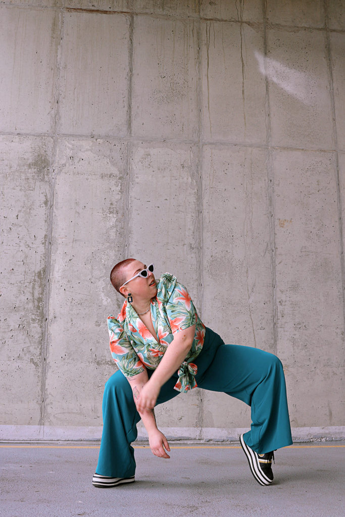 Becca Robinson, a woman with a buzzcut, wearing white-framed sunglasses, chunky
hoop earrings, a green and red Hawaiian shirt, turquoise pants, and black tennis shoes, poses in
front of a concrete wall. Her feet are wide apart with the heel of her left foot lifted. Her knees are
bent, and she is leaning to her right side, while looking upwards and to the left.