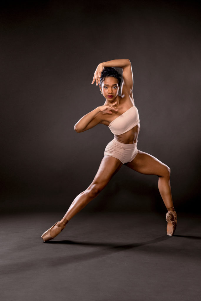 Dandara Veiga poses in a pale cropped tube top and matching briefs, wearing pointe shoes in a shade of bronze that matches her skin. She balances in a forced arch open fourth position, torso twisted toward the camera as she frames her face with her hands.