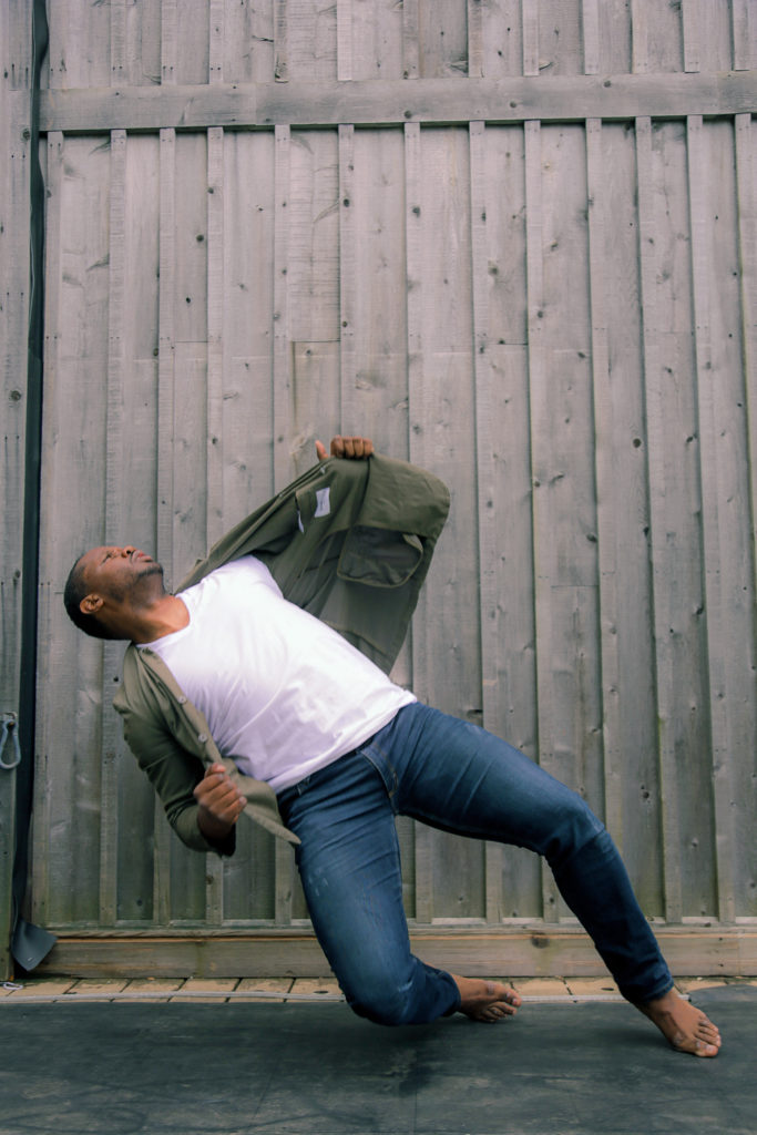 Dominic Moore-Dunson in blue jeans, white t-shirt, and green blazer dancing in front of a wooden wall.