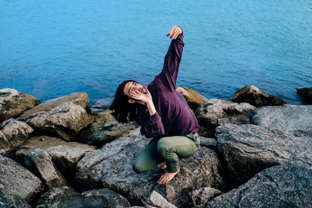 Elijah Richardson crouches on a series of boulders beside a body of water, long black hair flowing in the wind as he looks up toward an outstretched arm, fingers curling. His other hand rises near his mouth, somewhere between amplifying a call and shielding his face.