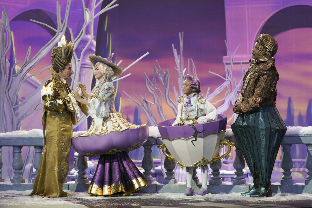 Four actors dressed as a fanciful candlestick, cups, and a clock stand in a line against a background of purple and white.