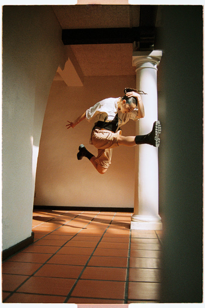 Gianna “Gigi” Todisco is jumping in the air, with one leg extended in front of her and the other bent behind her. One arm is wrapped around her head and the other is extended behind her. She is in the hallway of a white building with columns and a terra cotta colored tile floor. She is wearing black boots, cargo shorts, and a button up white shirt. Her dark hair is in braids. 