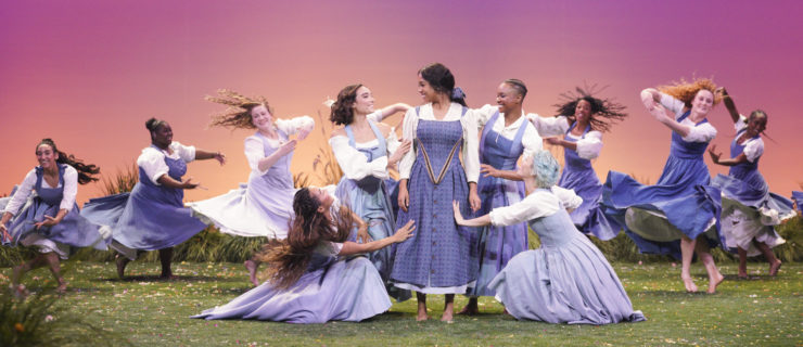 A dancer in a blue dress stands on a green field surrounded by dancers in blue dresses reaching out to her.