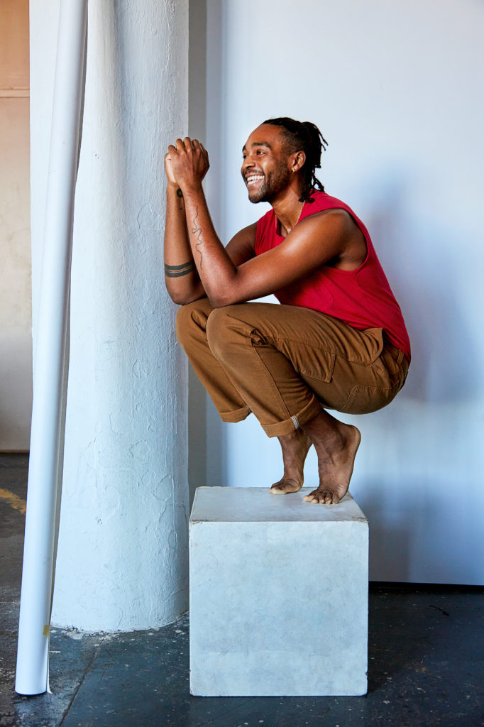 Jordan Demetrius Lloyd, a tall Black man crouched down on a white box. He’s smiling with his hands up.