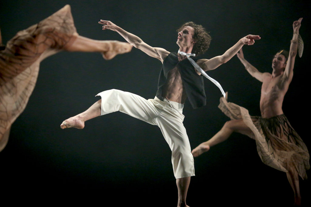 Mac Twining drifts through an off-kilter balance, arms floating up to shoulder height as one leg rises to a low side attitude. His hair fluffs out behind him as he directs his gaze on a upward diagonal. He wears short white trousers and a black vest open over a bare chest. Around him, male dancers in diaphanous skirts move through the same motion.