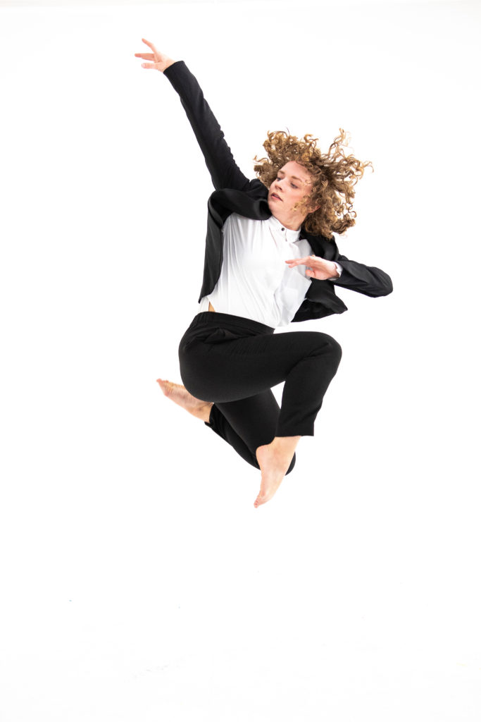 Madeline Maxine Gorman jumps in front of a white backdrop. Her knees are tucked up beneath her, feet pointed, while she twists to look toward the arm that is raised up and behind her. She wears a dark suit over a white button down. Her brown curls fly around her face.