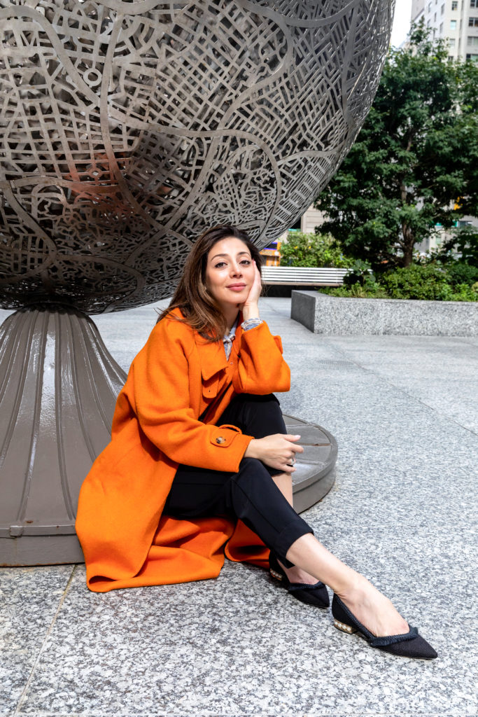 female wearing orange pea coat sitting by large round sculpture 