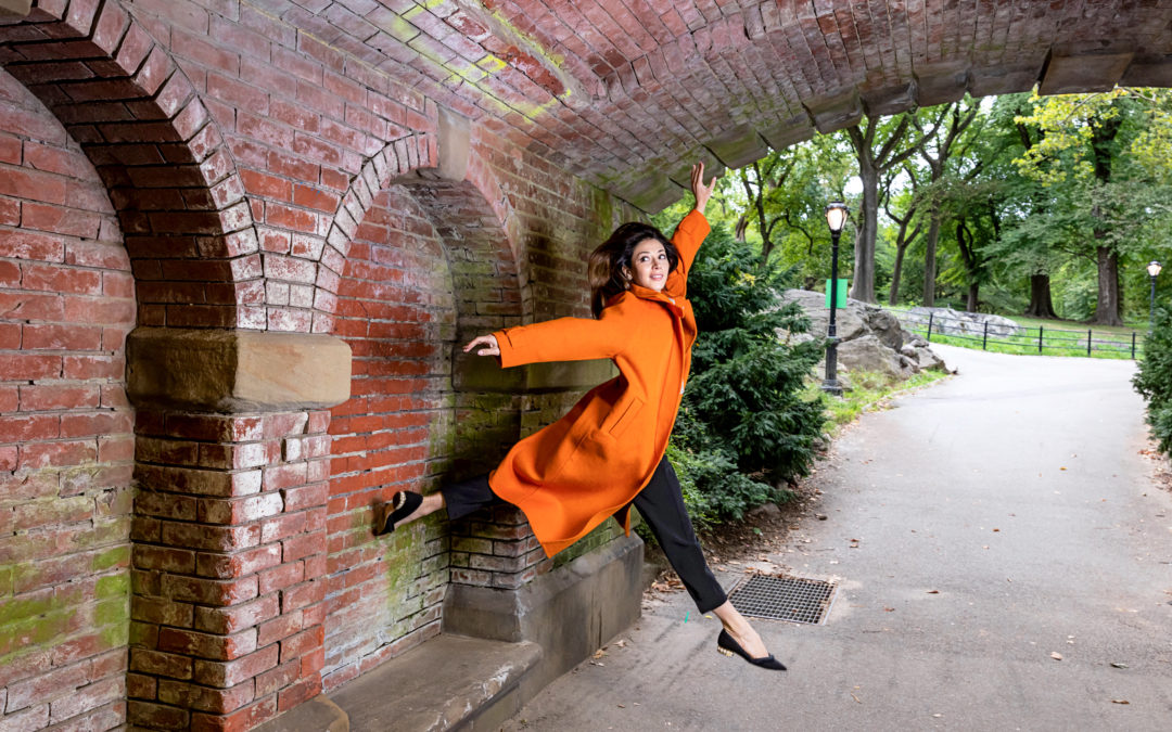 female dancer jumping under brick arch wearing an orange pea coat