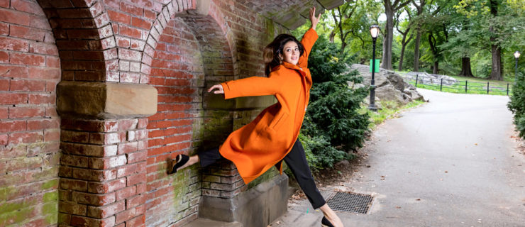 female dancer jumping under brick arch wearing an orange pea coat