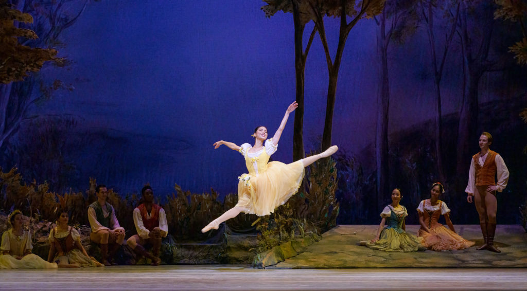 Mikaela Santos caught midair in a sissone, back arm raised on a diagonal to mirror her split legs. She smiles warmly, chin raised. She wears a yellow dress in the style of a romantic tutu. Around her other costumed dancers watch from the sides and back of the stage.