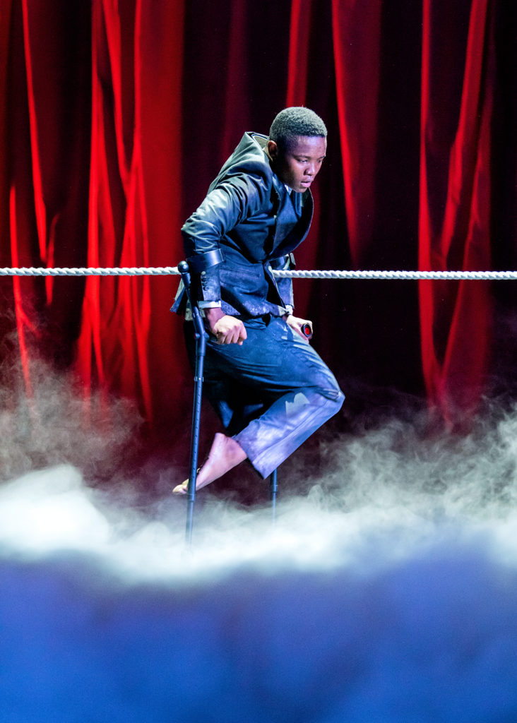 Musa Motha balances on his crutches, downstage leg bent at the knee with a pointed foot. He hovers over a sea of fog against the backdrop of a red velvet curtain. A rope at the height of his waist runs out of frame to each side. His gaze is meditative as he peers down into the fog.