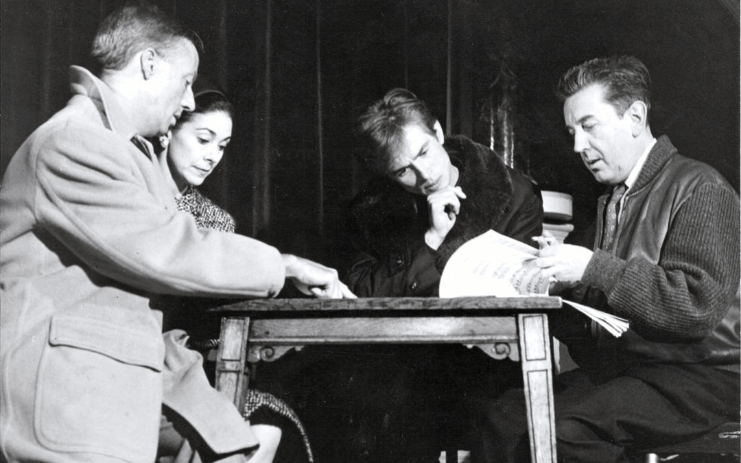 In a black and white archival photo, three men and one woman sit at a small table. Sir Frederick Ashton turns the pages of a musical score while Rudolf Nureyev brings his hand to his chin, frowning over it. Margot Fonteyn looks down at what William Chappell is pointing to on the table with one hand.