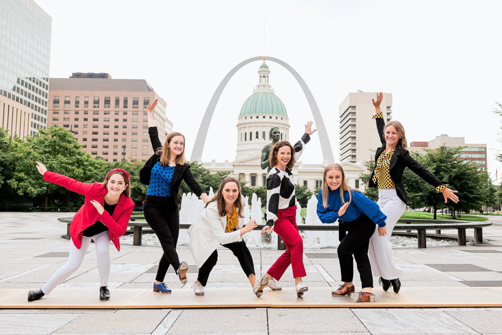 A half dozen smiling women in tap shoes pose on a tap board in front of the Gateway Arch in St. Louis.