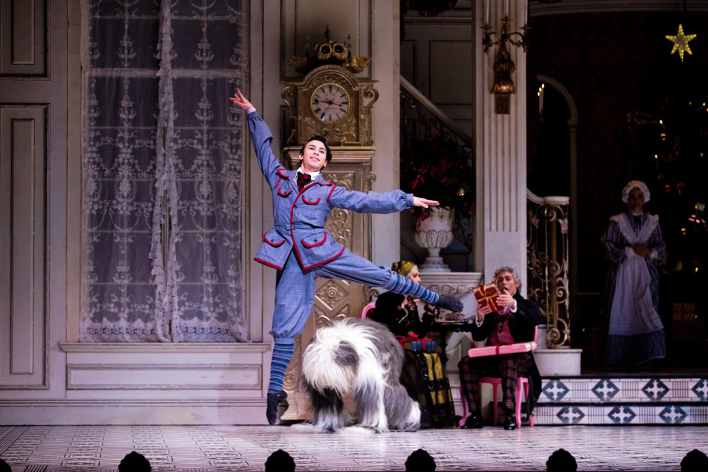 Simone Acri is midair, doing a temps levé. He is costumed in an old-fashioned, childlike blue suit with red piping. A dancer costumed as a shaggy dog appears behind him, seeming ready to pounce.