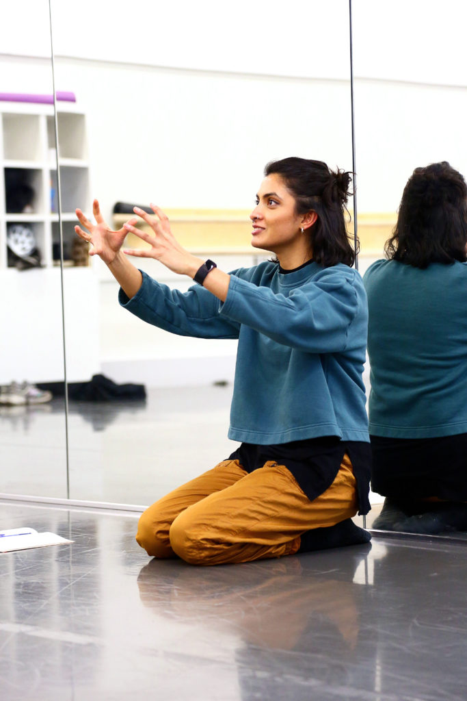 Vidya Patel kneels at the front of a studio, an excited smile on her face as she gestures with her arms in front of her as though holding an invisible ball.