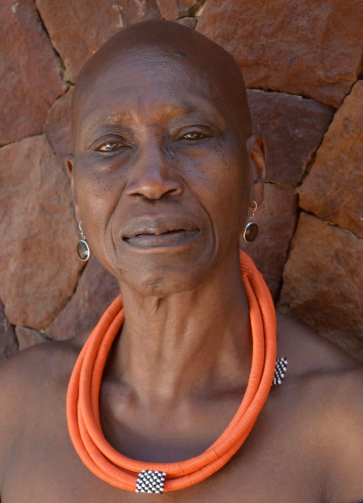 A portrait of Germaine Acogny. She is a dark-skinned, older woman with a shaved head and light brown eyes. She stands in front of a red and orange stone wall, gazing seriously at the camera. An orange, corded necklace is looped around her neck.