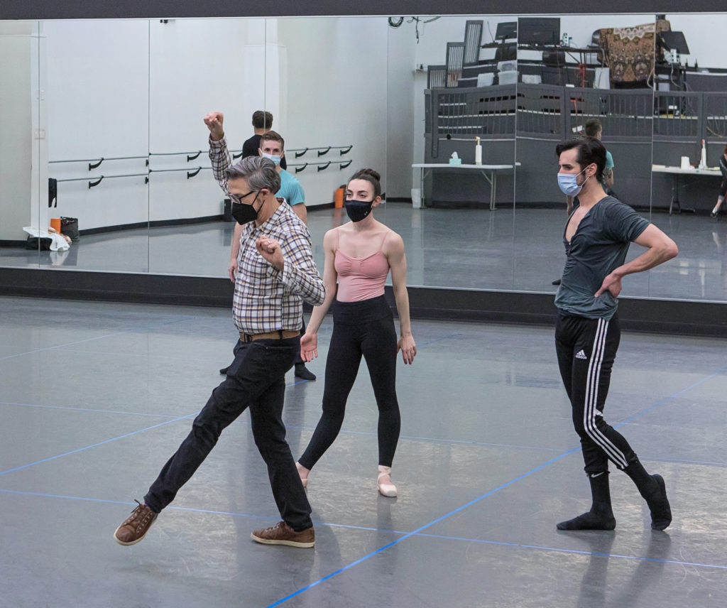 In a rehearsal studio, Paul Vasterling brushes a foot forward, gaze down as he explains a note to the pair of dancers watching behind him. All wear face masks.