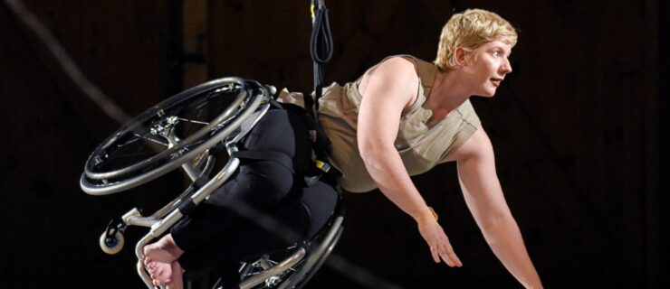 A white dancer with cropped blonde hair is suspended midair. She looks to the right, arms extended from pushing off the ground, body and wheelchair diagonal to the floor. A black cord at her waist extends up.