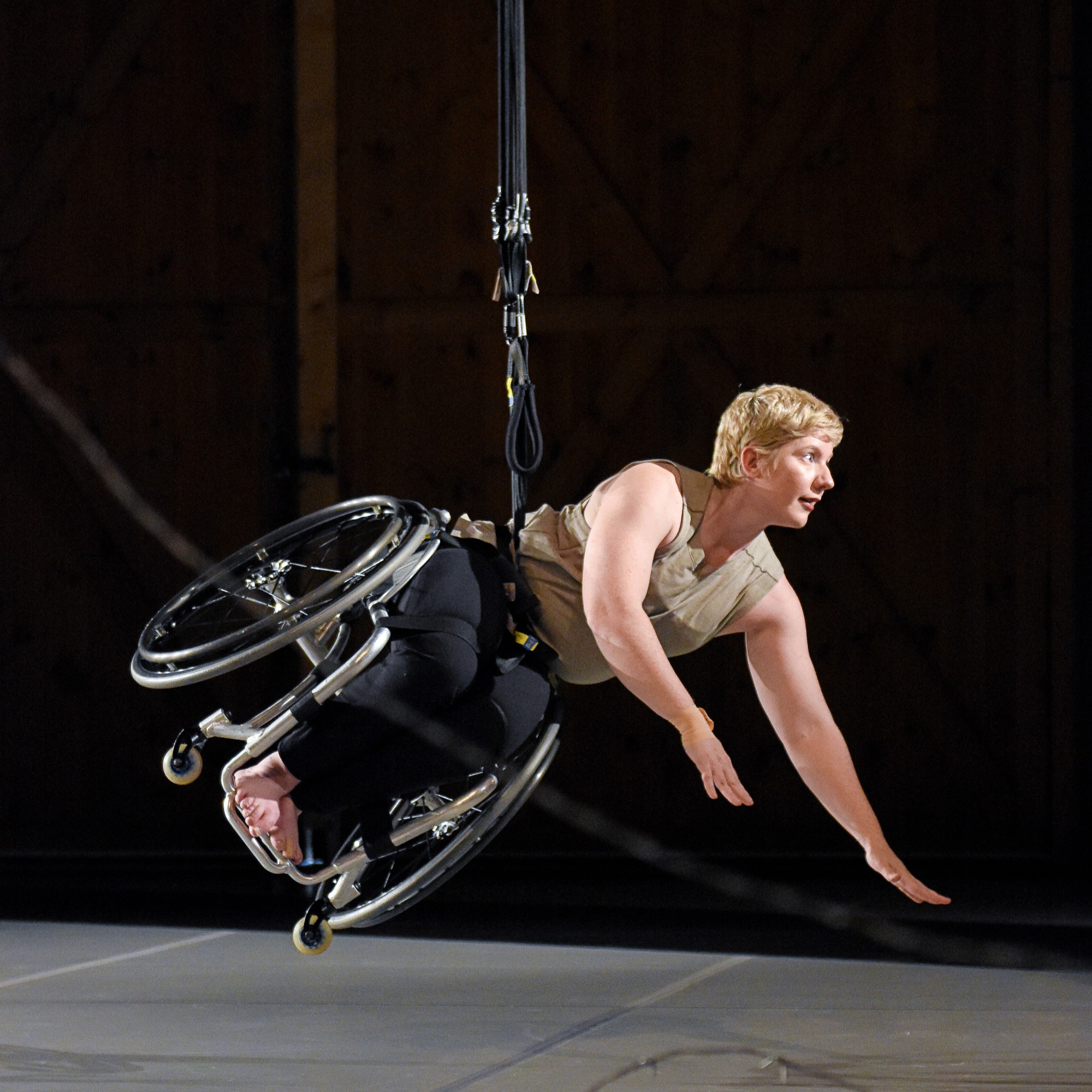 A white dancer with cropped blonde hair is suspended midair. She looks to the right, arms extended from pushing off the ground, body and wheelchair diagonal to the floor. A black cord at her waist extends up.