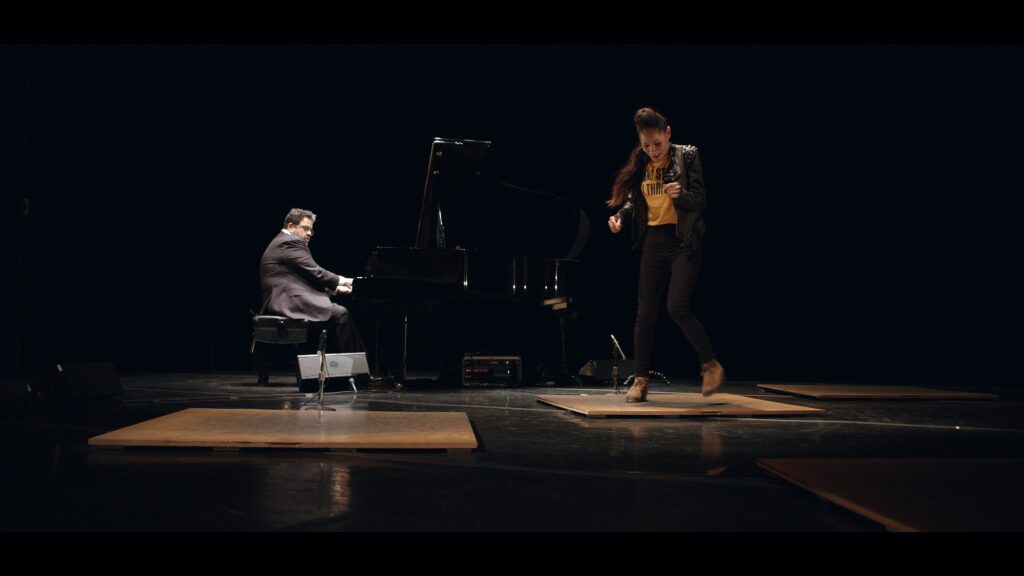 Ayodele Casel looks towards her feet, which are partially blurred as she taps on a square tap board. Upstage and to her right, a besuited pianist looks past the piano to her, his hands on the keys.