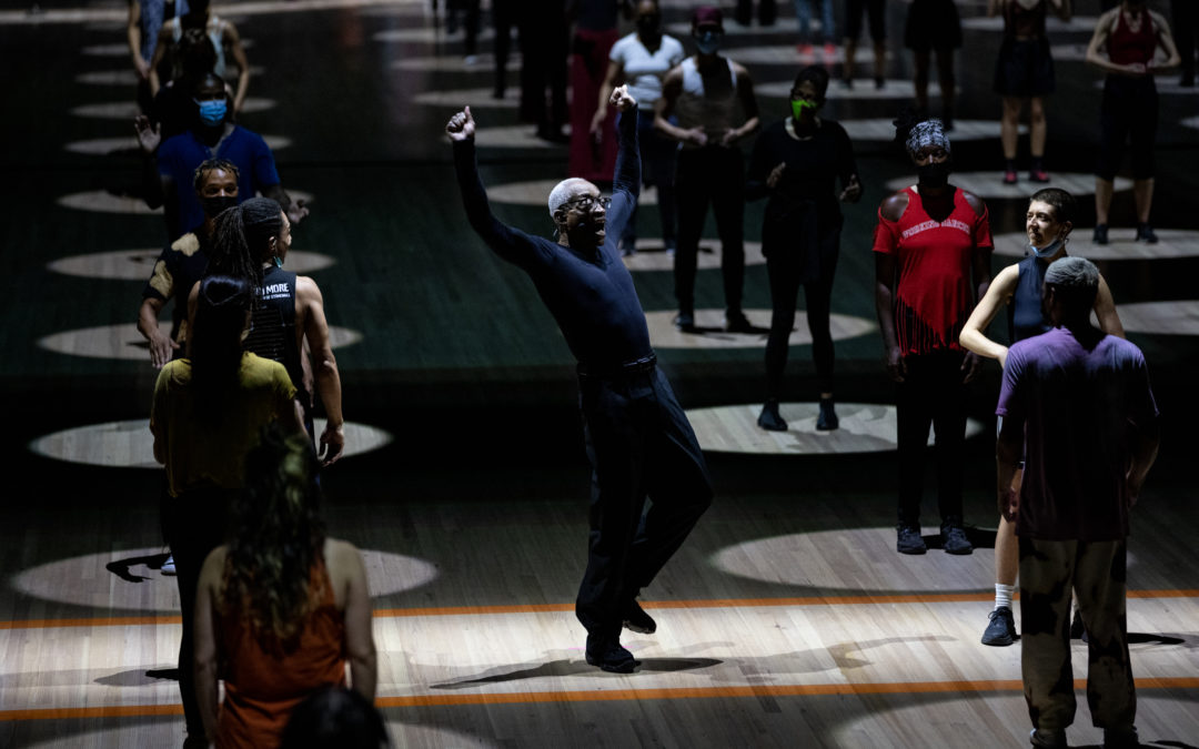 Bill T. Jones, an older Black man, balances with one flexed foot raised slightly off the ground. His arms rise beside his head as his torso leans back. Around him are lines of audience members standing in evenly spaced spotlights.