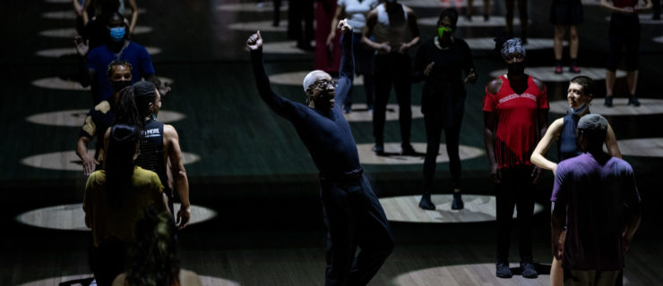 Bill T. Jones, an older Black man, balances with one flexed foot raised slightly off the ground. His arms rise beside his head as his torso leans back. Around him are lines of audience members standing in evenly spaced spotlights.