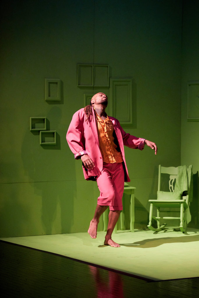 A dancer clothed in bright pink and orange closes his eyes as he tips his head back, grooving alone on a set designed to evoke a small apartment painted entirely in lime green.